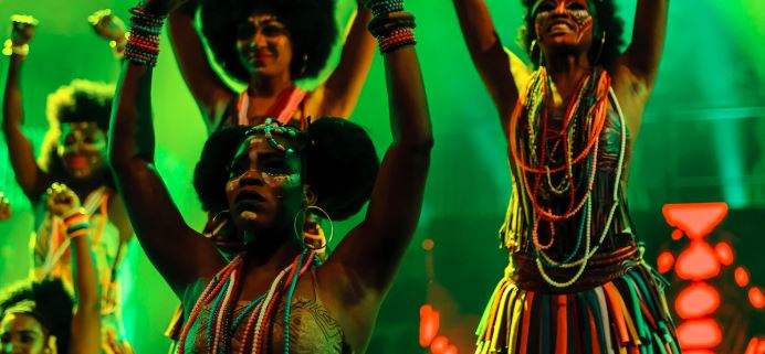 Stage still showing a live Afrobeat band with three Queens in adire costumes performing Yoruba‑inspired choreography for the Kalakuta Queens stage play.